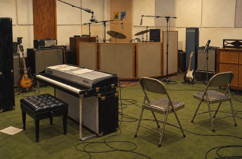 A vintage recording studio: an electric piano and bench in the foreground, two folding chairs facing each other, guitars and amplifiers around the room